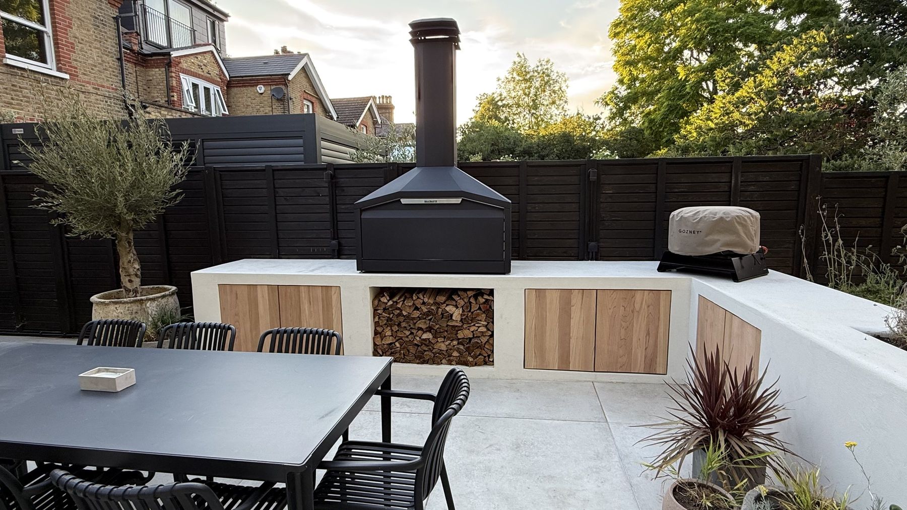 Rendered outdoor kitchen with a black log-burning oven, timber cupboard doors and a log store, built by Oakdene Builders